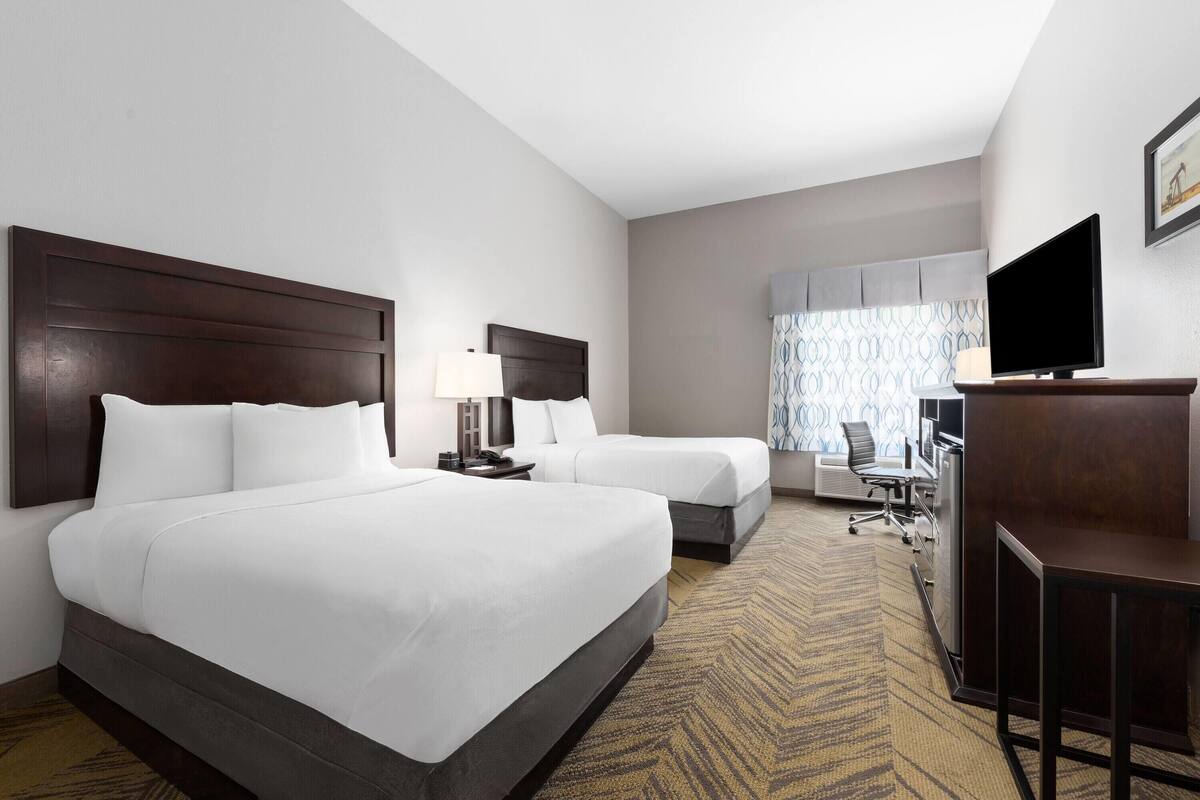 A hotel room with two beds, a desk, a chair, a television, and a window with patterned curtains.