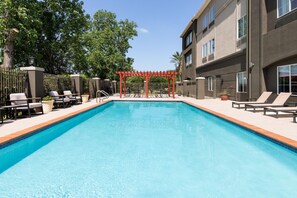 Una piscina rectangular con una pérgola roja en el centro, rodeada de sillas de descanso y una valla.