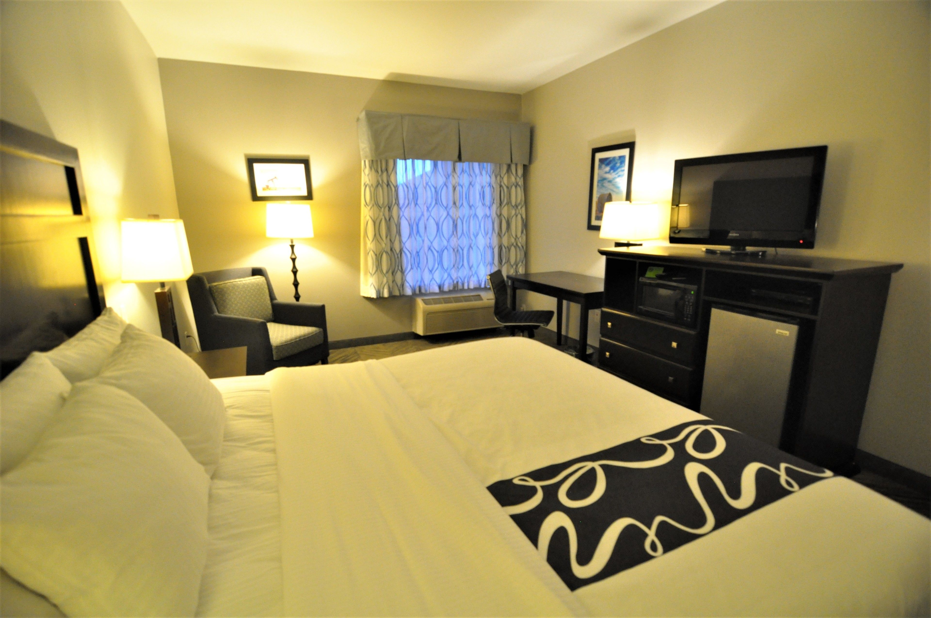 A hotel room with a large bed, a desk with a television, a chair, and a lamp.