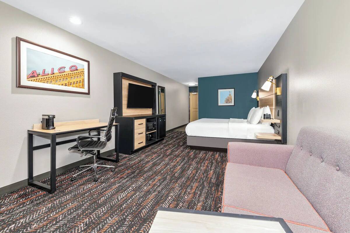 Una habitación de hotel con una cama, un escritorio con una silla, un televisor, una cafetera y un cuadro en la pared.
