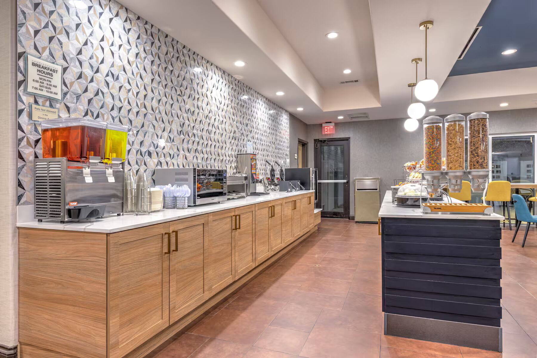 A modern breakfast area with a geometric patterned wall, a counter with beverage dispensers, and a snack station.