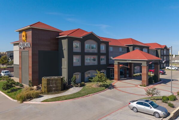A multi-story hotel with a red roof, a prominent logo on the facade, and a parking area with cars.