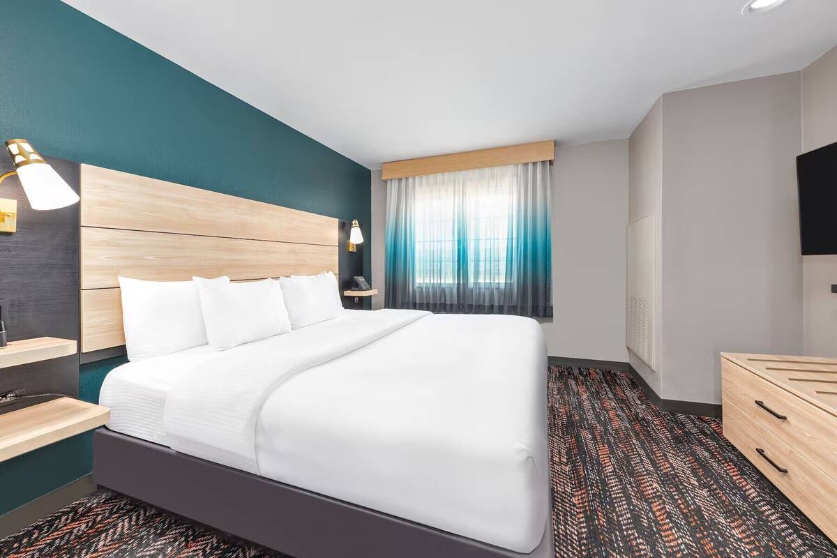 Habitación de hotel con una cama grande, una mesita de noche, un televisor y una ventana con cortinas.