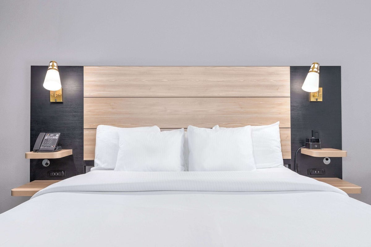 Una habitación de hotel moderna con cabecera de madera, ropa de cama blanca y mesitas de noche con lámparas y teléfono.
