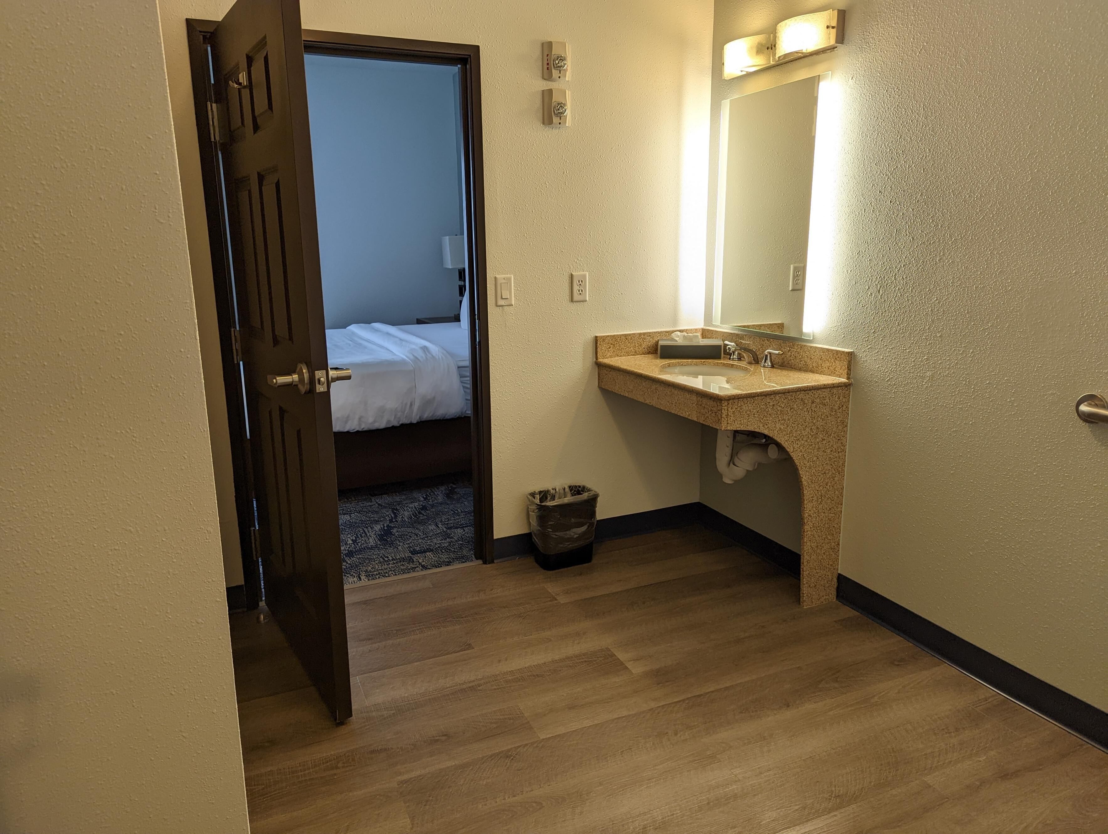 A hotel room with a wooden floor, a bed with white bedding, a sink with a mirror, and a trash bin.
