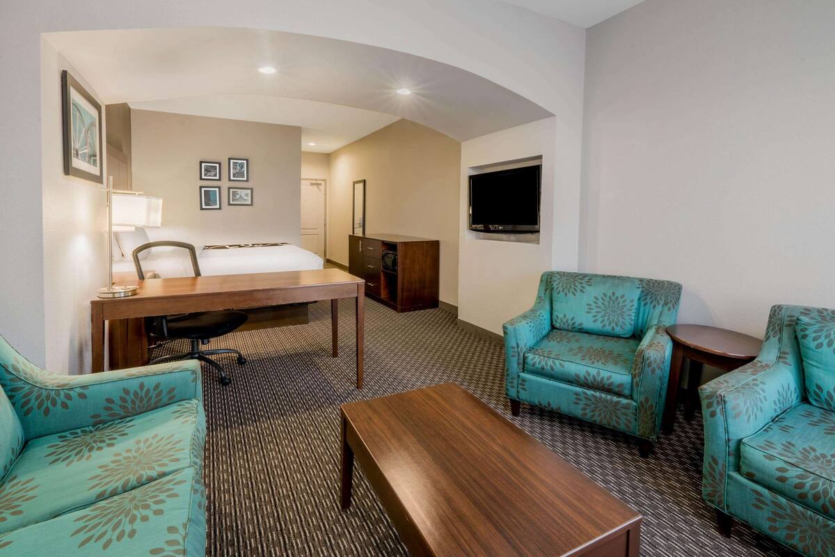 Een hotelkamer met een bed, twee armstoelen met een blauwgroen patroon, een houten salontafel, een bureau met een lamp, een flatscreen televisie aan de muur en een vloerbedekking.