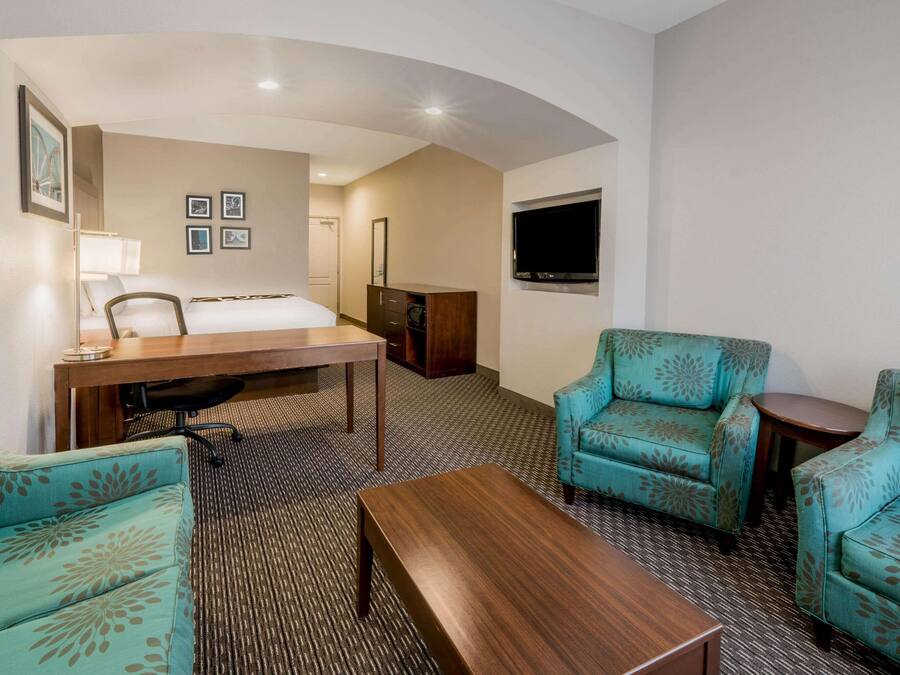 A hotel room with a bed, two teal patterned armchairs, a wooden coffee table, a desk with a lamp, a flat-screen TV mounted on the wall, and a carpeted floor.