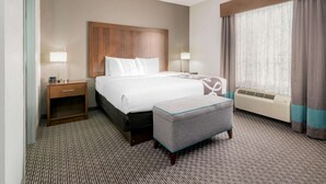 A hotel room with a bed, bedside tables, an ottoman, a window with curtains, and a wall-mounted heater.