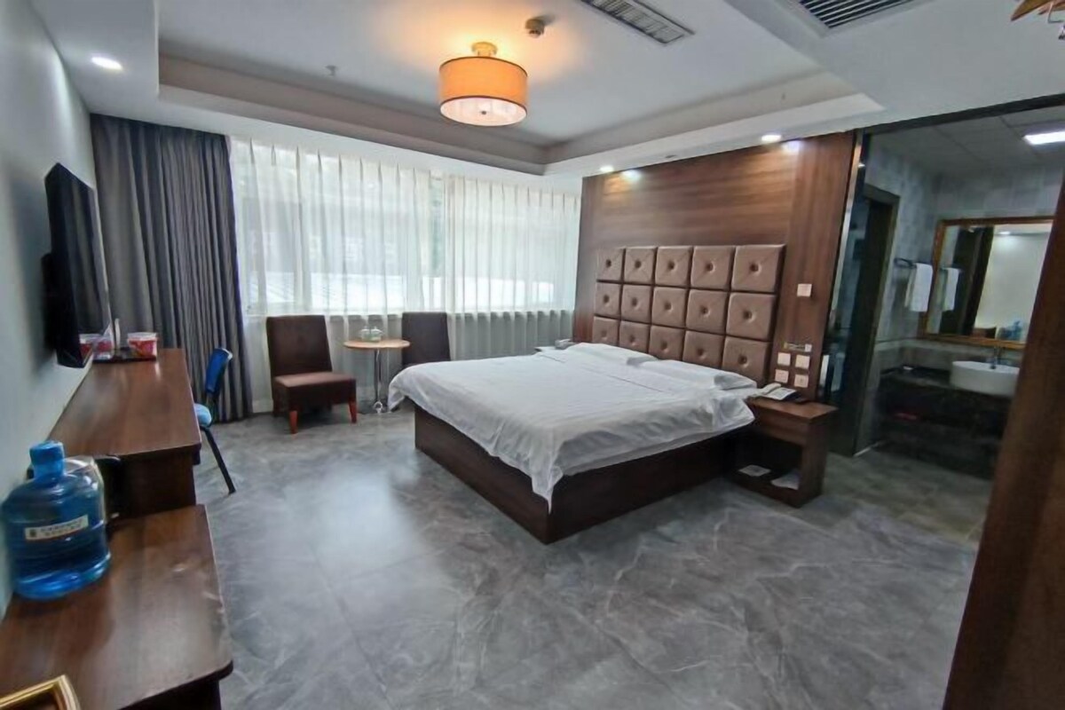 A hotel room with a large bed, a wooden headboard, a desk with a chair, a large water dispenser, and a bathroom with a sink and mirror.