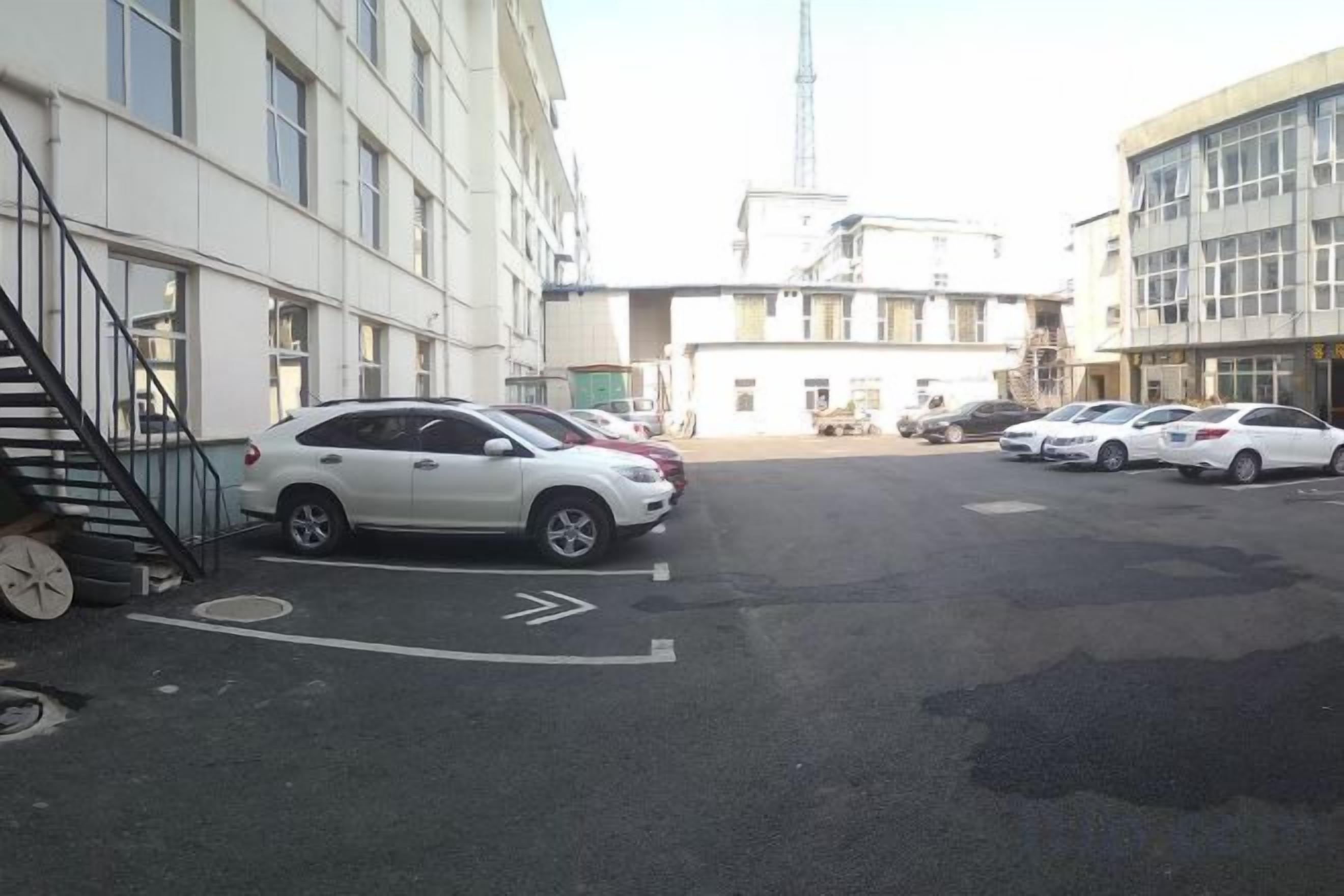 Un estacionamiento con varios coches, un edificio con una escalera a la izquierda y otro edificio con una torre al fondo.