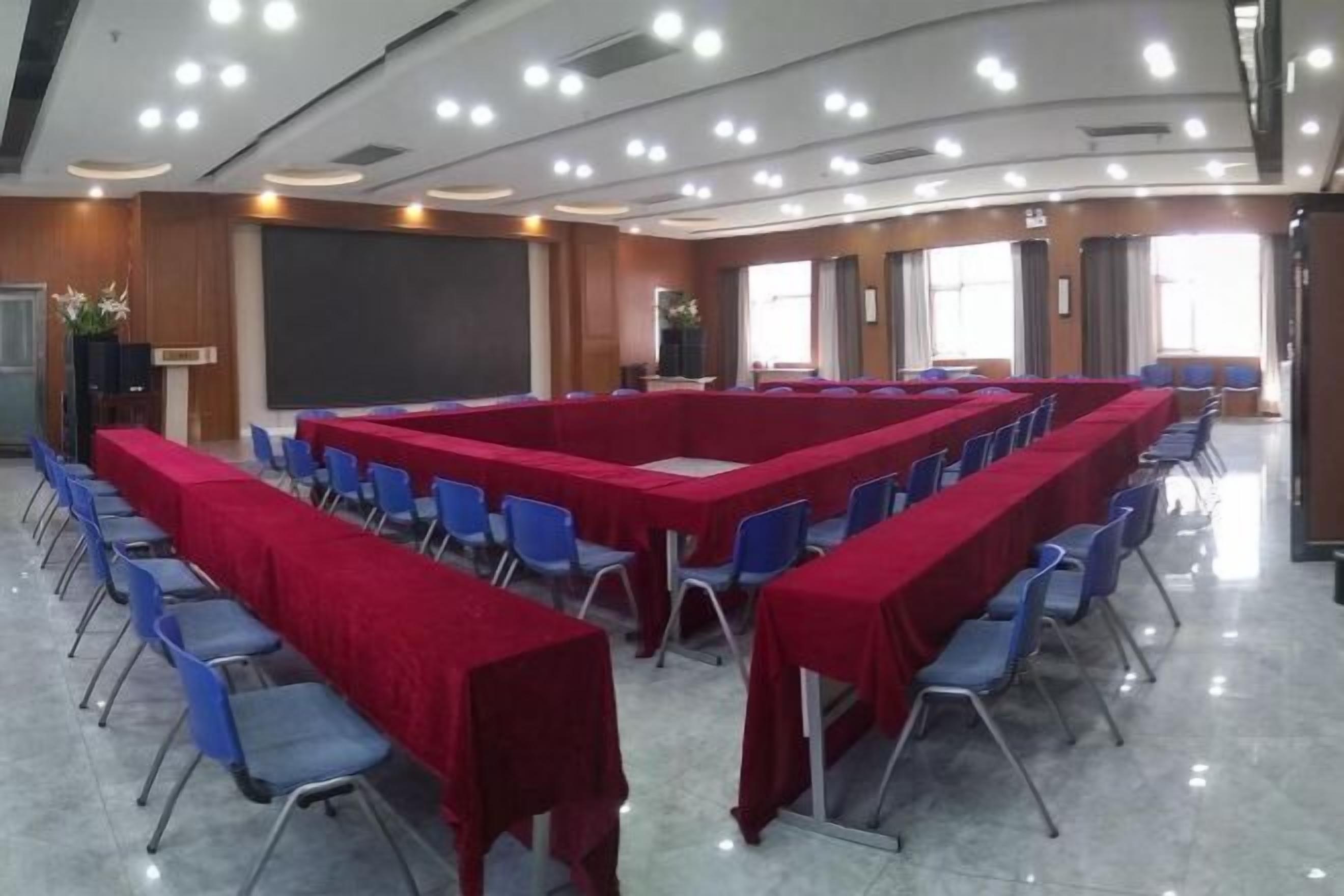 A conference room with a long table covered in red, surrounded by blue chairs, and a large screen at the front.