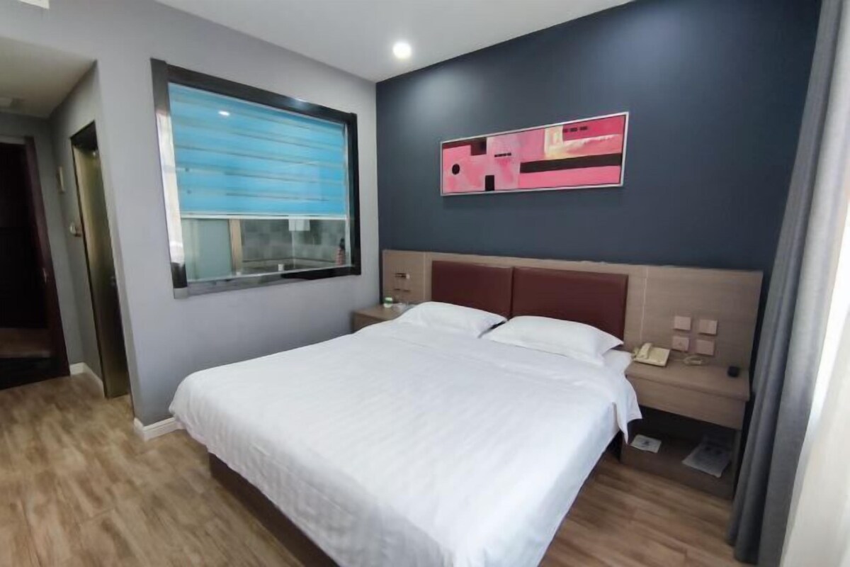 A hotel room with a bed, a nightstand, a window with blinds, and a wall-mounted artwork.