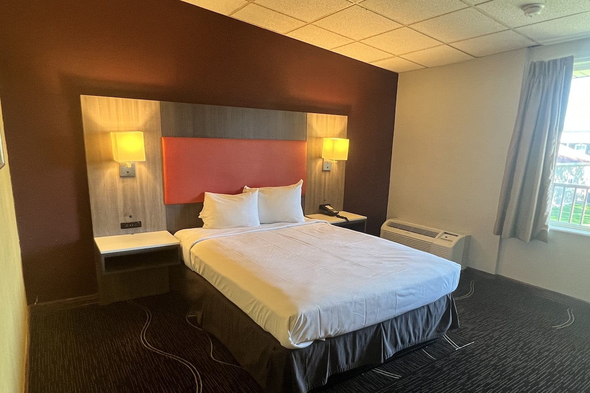 A hotel room with a large bed, two bedside tables, a desk, a chair, a lamp, and a window with curtains.