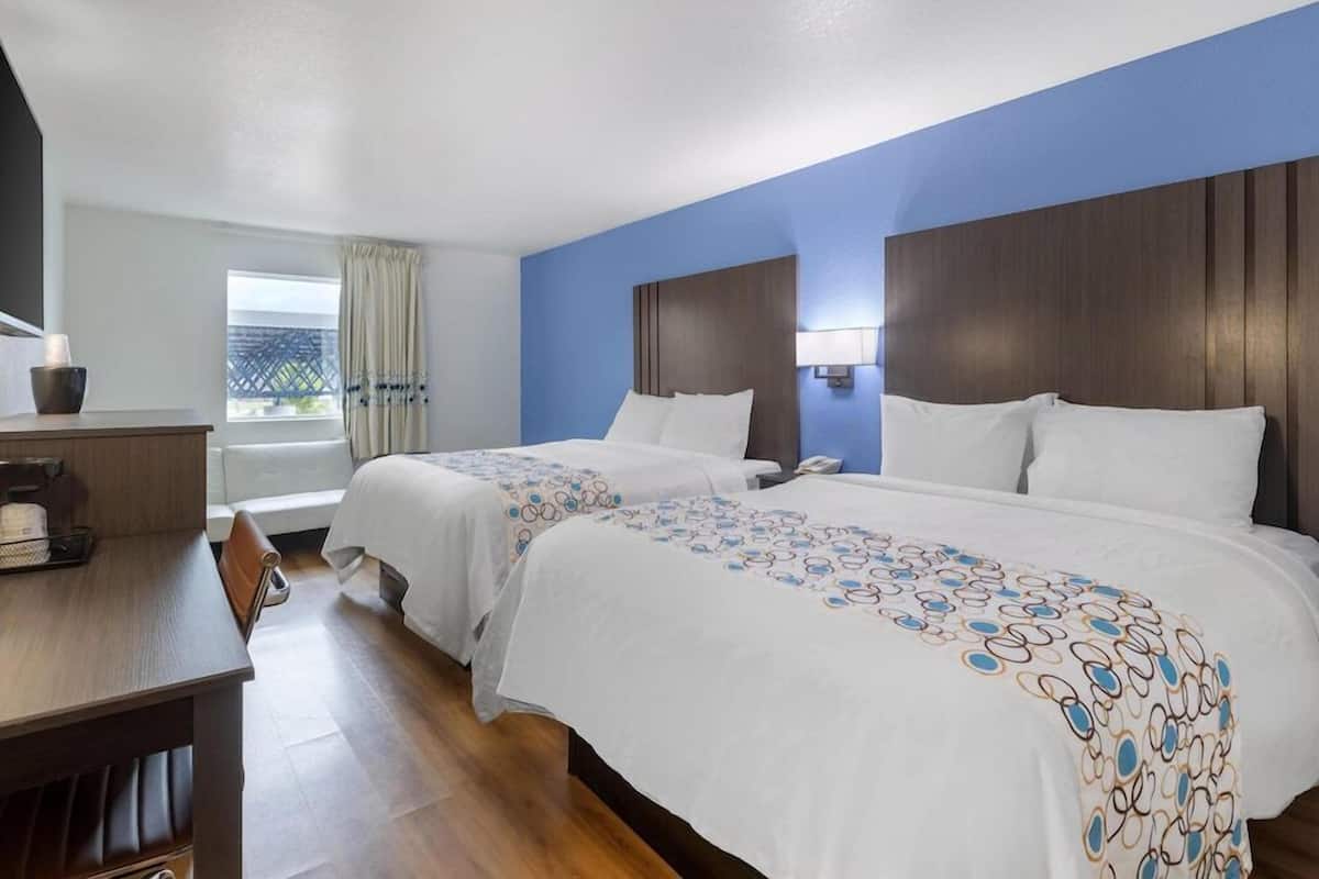Une chambre d’hôtel avec deux lits, un bureau, une chaise, une fenêtre avec des rideaux et une vue sur un bâtiment à l’extérieur.