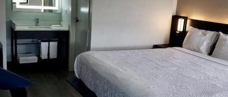 A hotel room with a bed, a bedside table, a nightstand, a mirror, and a bathroom with a sink and towel rack.