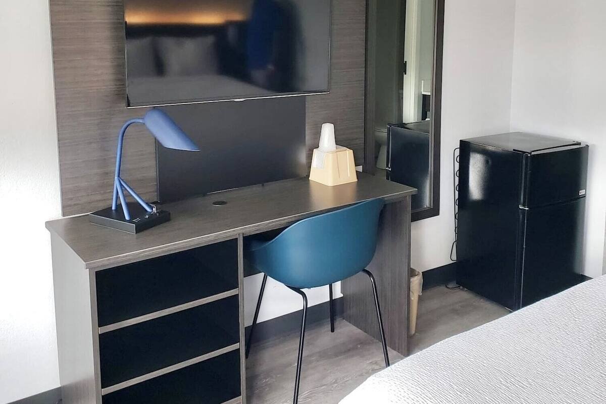 A modern hotel room with a flat-screen TV mounted on a wall, a desk with a chair, a small refrigerator, and a bed with a textured blanket.