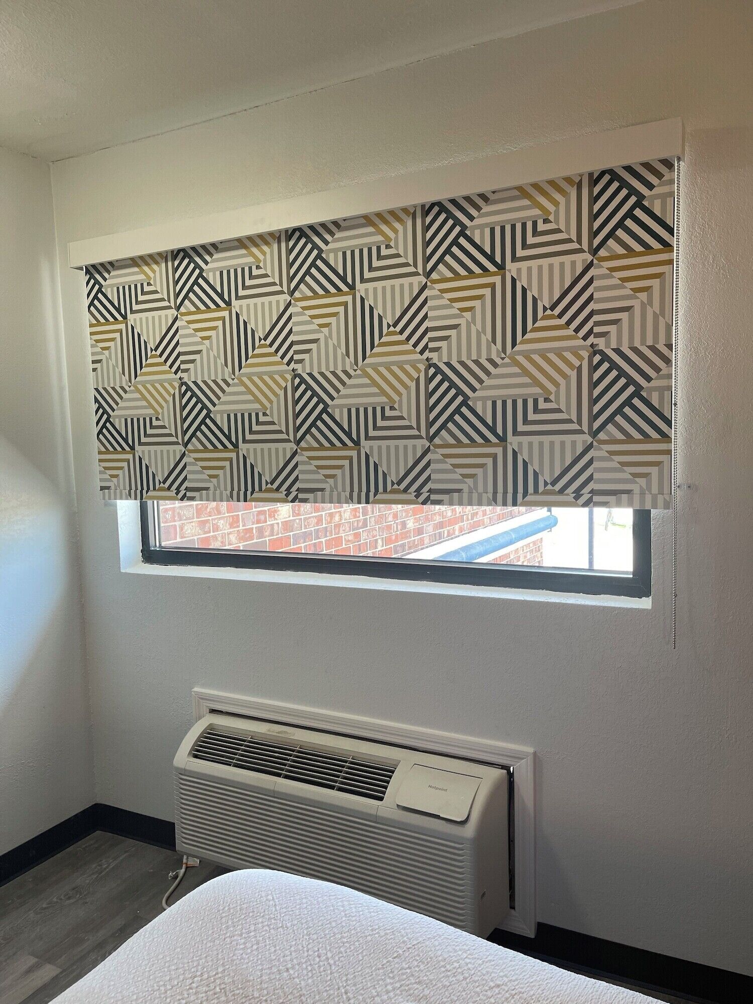 A room with a window covered by a patterned roller blind, a radiator below the window, and a visible brick wall outside.
