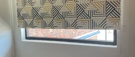 A room with a window covered by a patterned roller blind, a radiator below the window, and a visible brick wall outside.