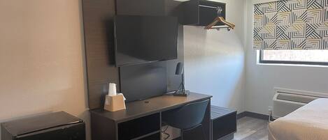 A compact hotel room with a bed, a desk with a chair, a television, and a radiator.