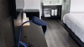 A compact hotel room with a bed, a desk with a computer, a TV, a bathroom with a mirror, and a window.
