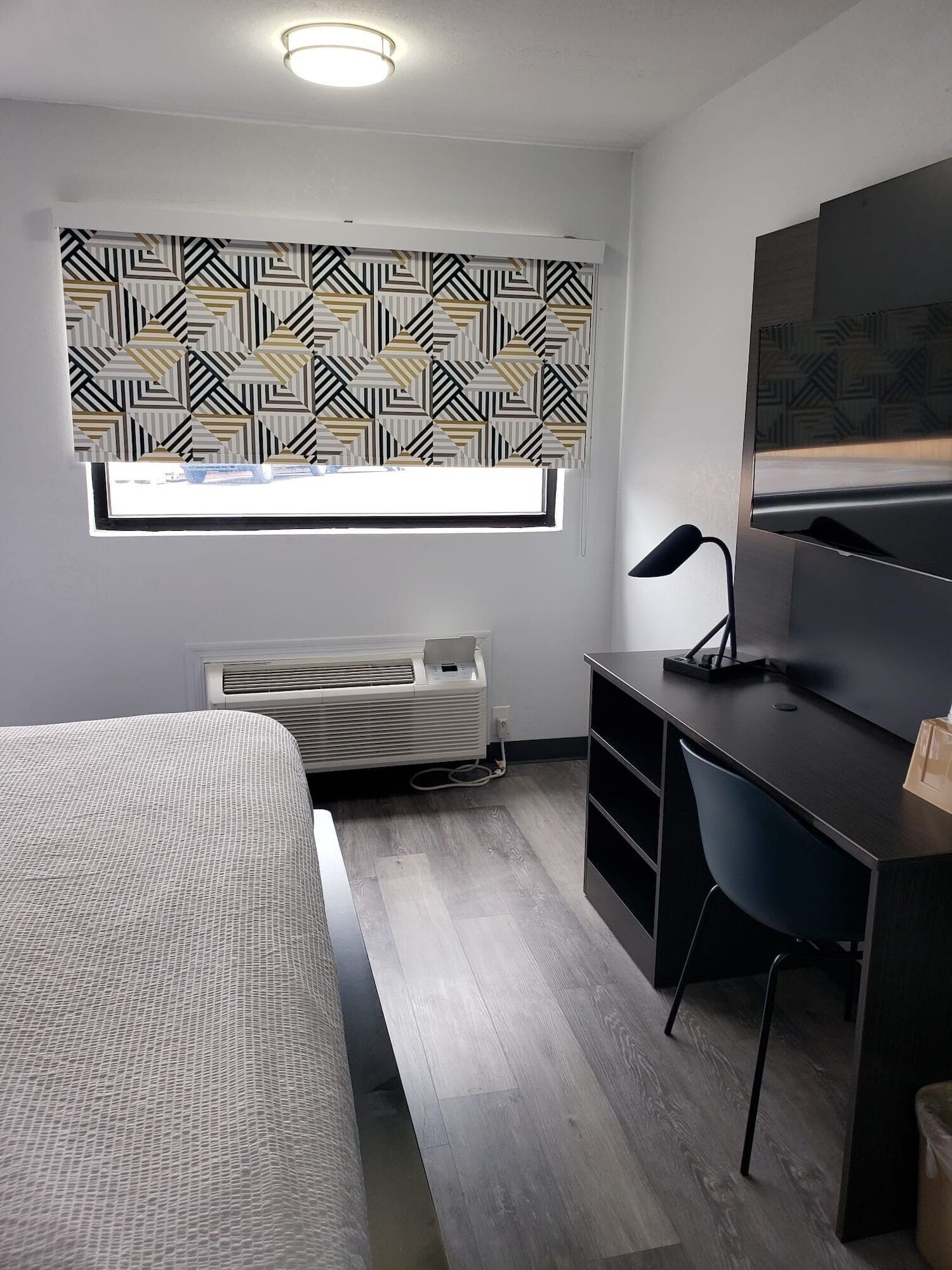 A modern hotel room with a bed, a desk, a chair, a large window with patterned curtains, a ceiling light, and a wall-mounted TV.