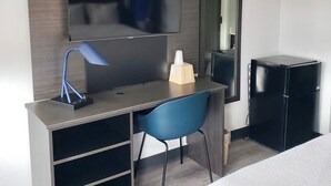 Une chambre d’hôtel moderne dotée d’un téléviseur à écran plat fixé à un mur en bois, d’un bureau avec une chaise, d’un petit réfrigérateur et d’un lit recouvert d’une couverture à motifs.