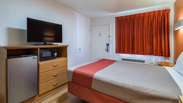 A compact hotel room with a bed, a TV, a minibar, and a door with a window.