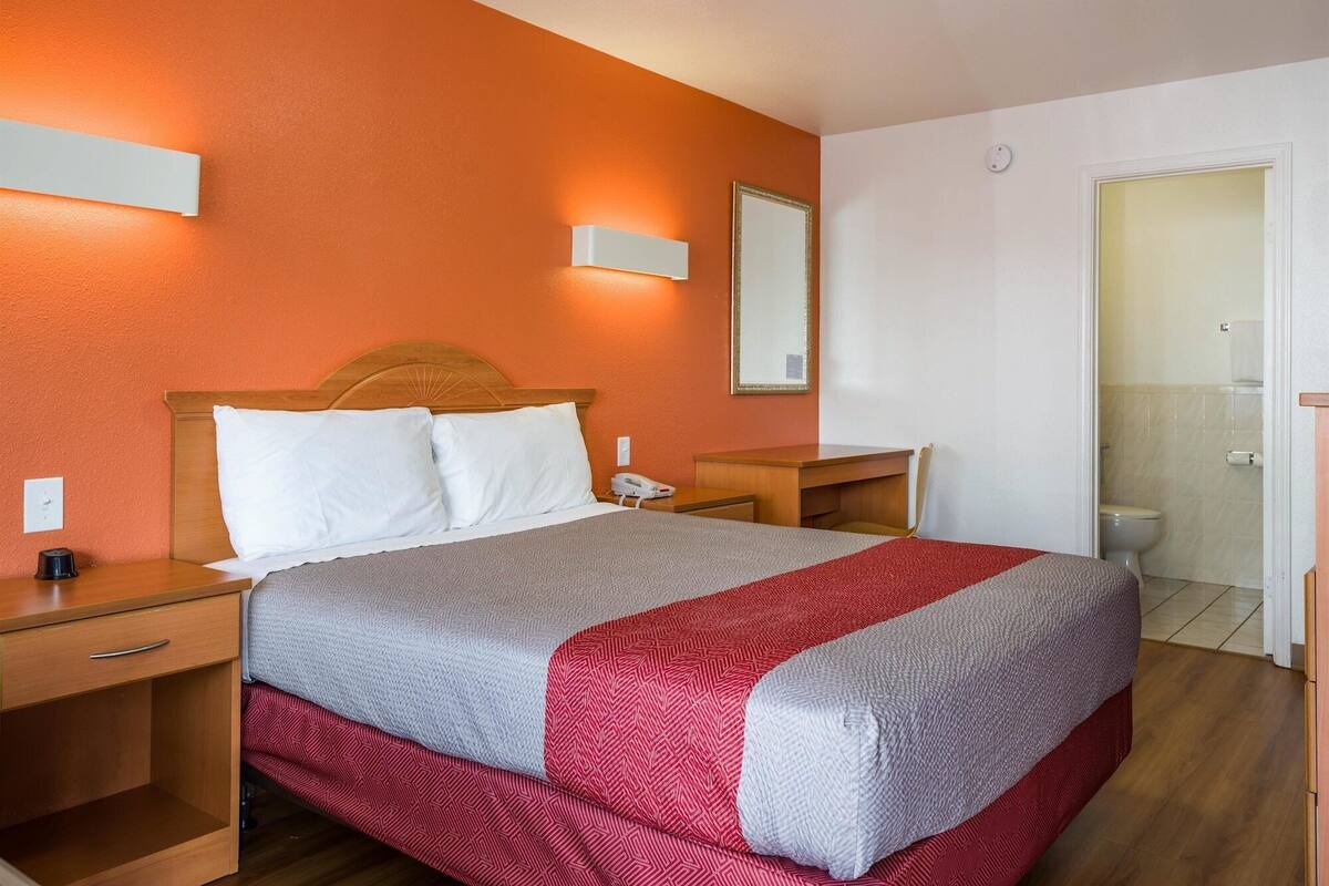 A hotel room with a wooden bed, a bedside table with a phone, a nightstand, and a bathroom visible through an open door.