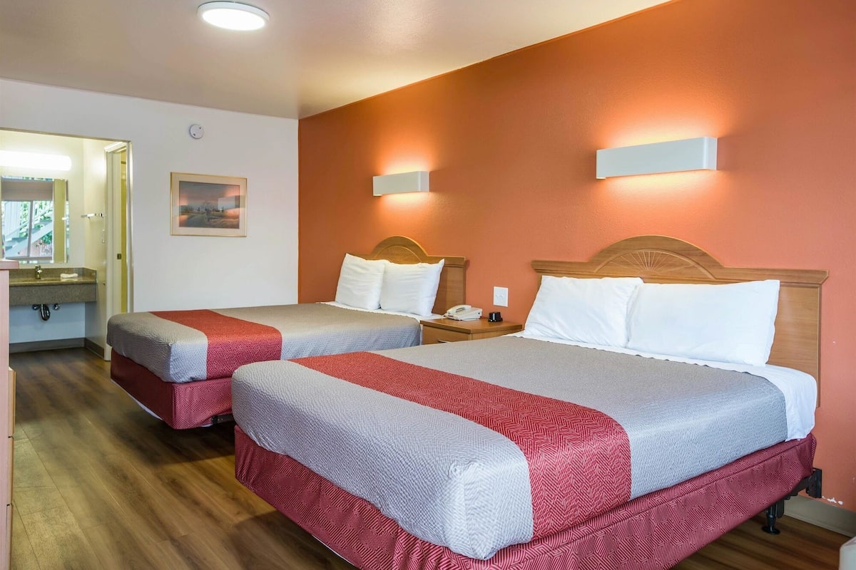 A hotel room with two beds, a wooden headboard, a nightstand, a bedside table, and a wall-mounted light fixture.