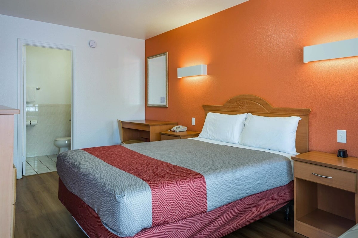 A hotel room with a bed, bedside tables, a nightstand, a mirror, and a bathroom visible through an open door.