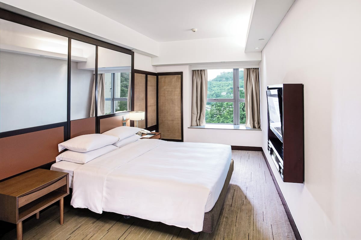 Une chambre d’hôtel avec un grand lit, une table de chevet, une télévision et une fenêtre donnant sur un paysage verdoyant.