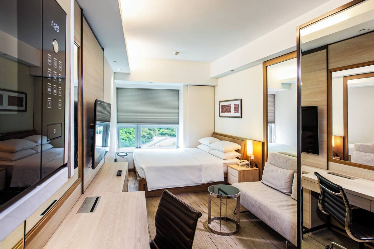 Une chambre d’hôtel équipée d’un lit, d’un bureau, d’une chaise, d’une télévision, d’un miroir et d’une fenêtre donnant sur un paysage verdoyant.
