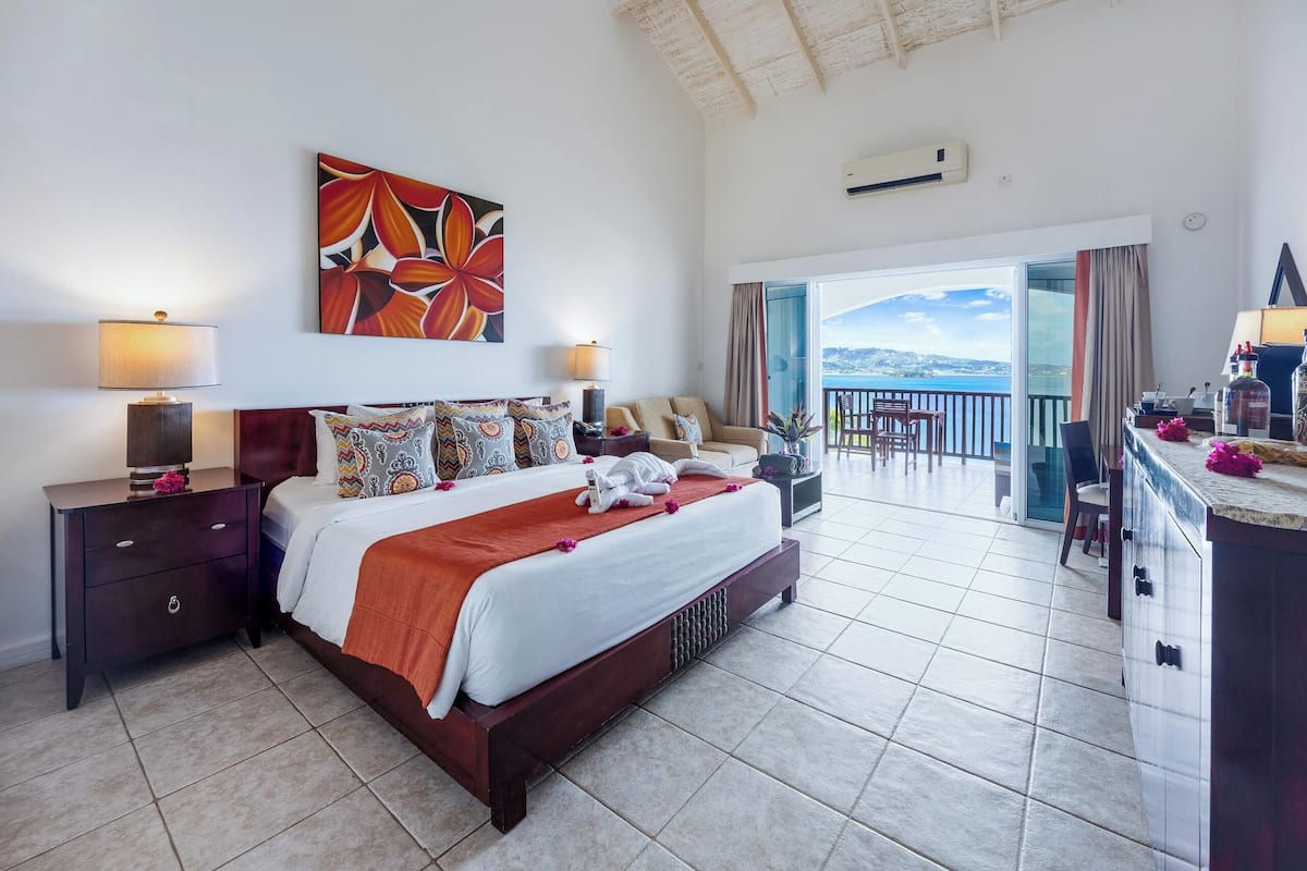 A hotel room with a large bed, two bedside tables, a painting, and a balcony with a view.