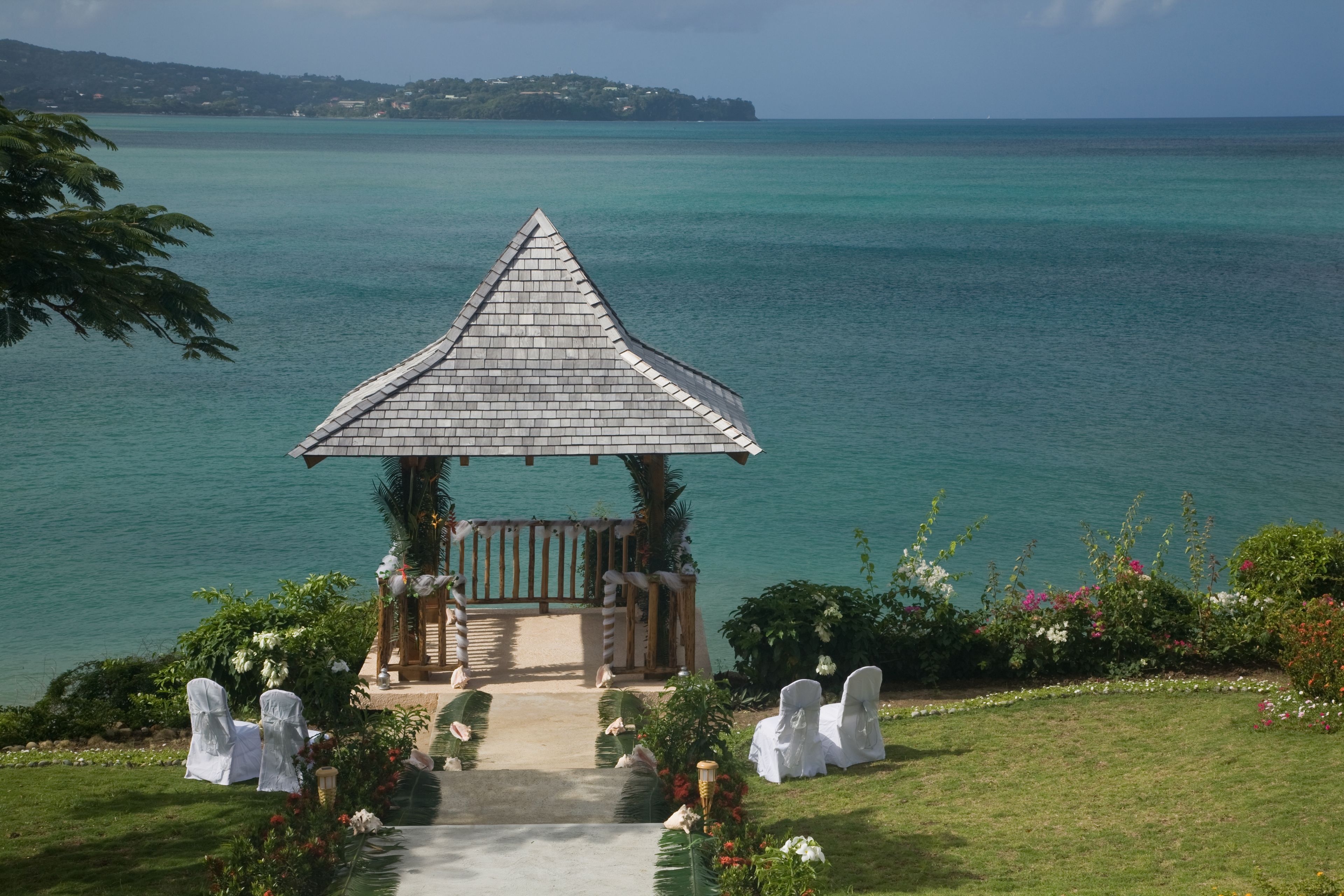A gazebo with a tiled roof overlooking a body of water, surrounded by greenery and white chairs.