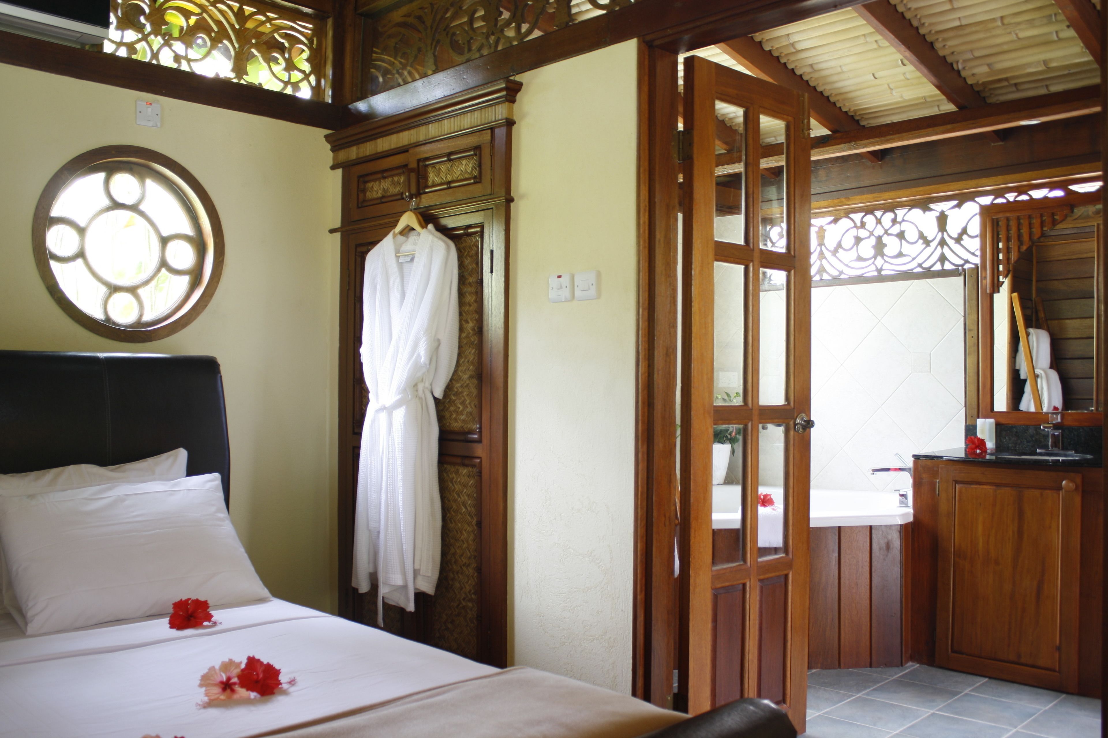A hotel room with a bed, a robe hanging on a door, a bathroom with a sink and mirror, and a decorative window.
