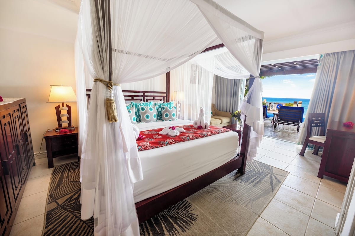 A hotel room with a four-poster bed, a desk with a lamp, and a view of the ocean.