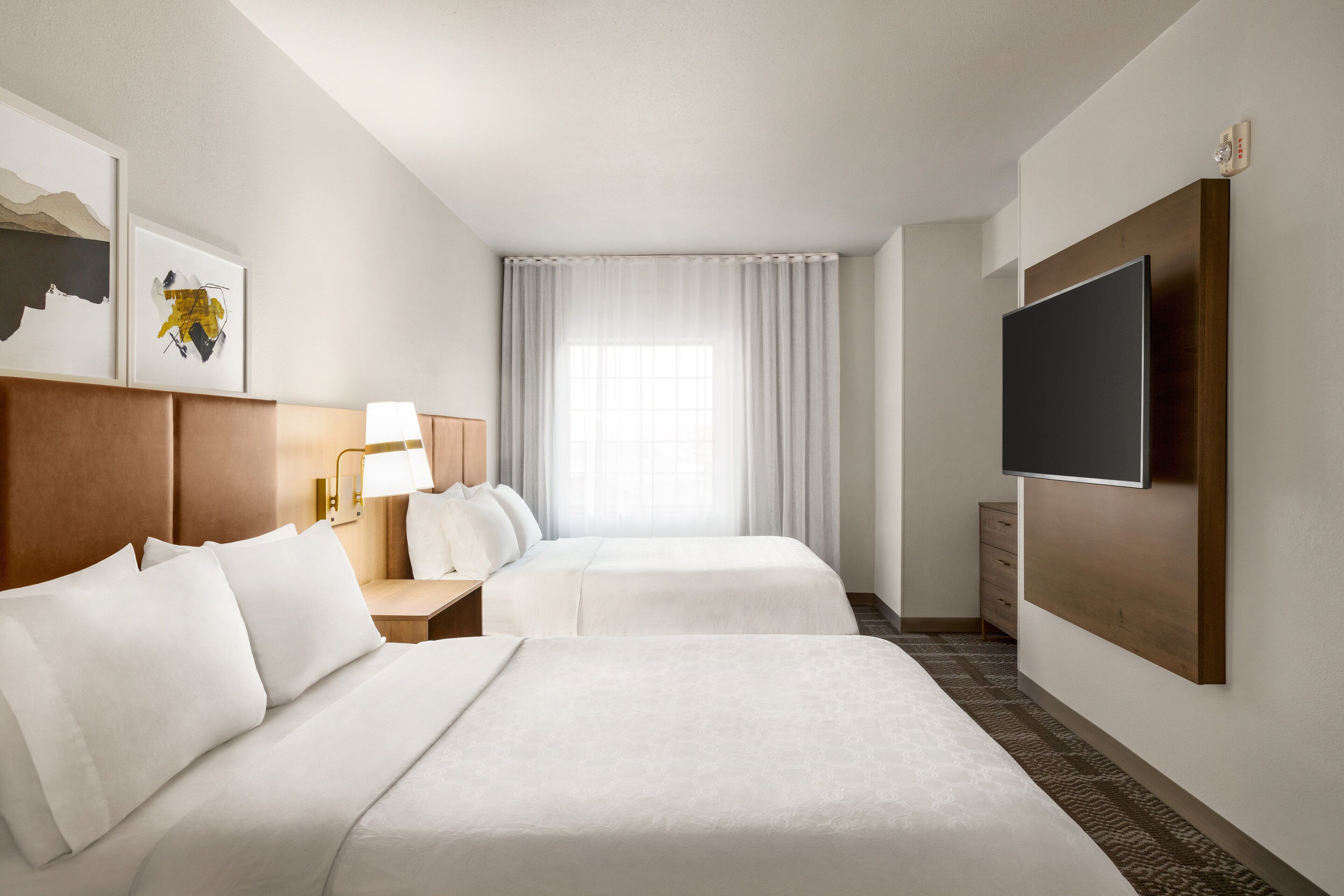 A hotel room with two beds, a flat-screen TV, a desk with a lamp, and a window with curtains.