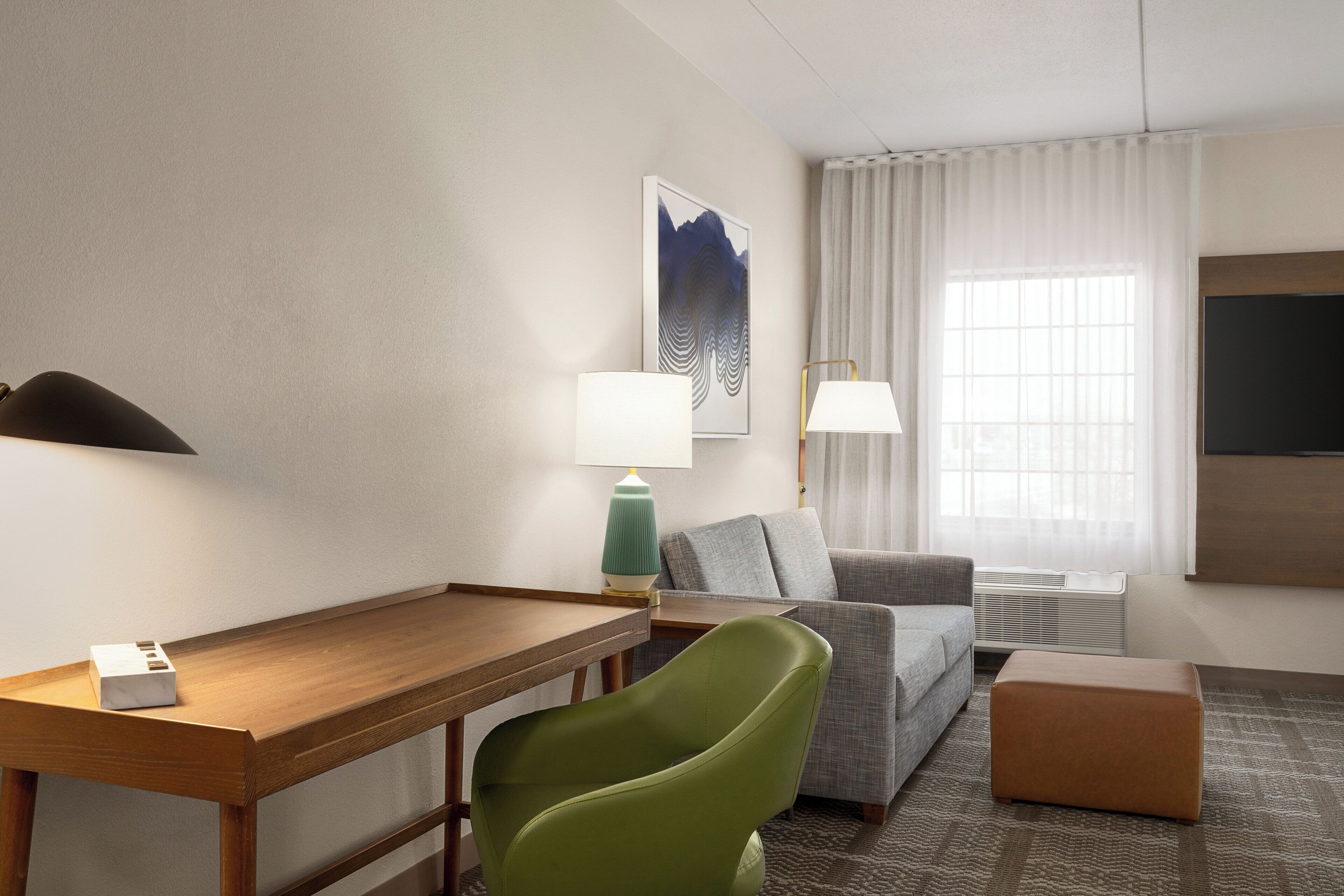 A hotel room with a wooden desk, a green chair, a grey sofa, a small ottoman, a flat-screen TV, and a window with curtains.