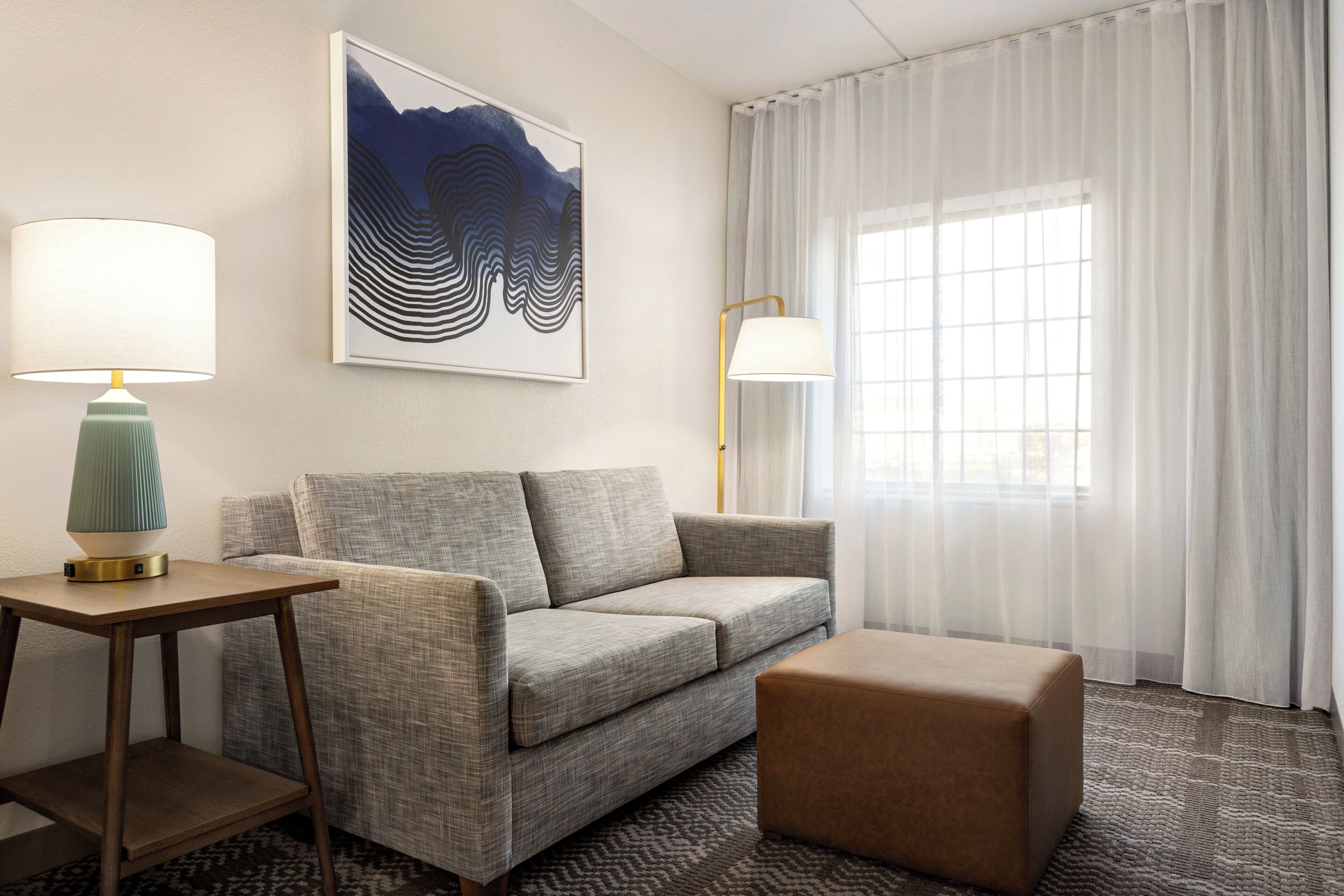 A hotel room with a sofa, a small table with a lamp, a large window with sheer curtains, and a framed artwork on the wall.