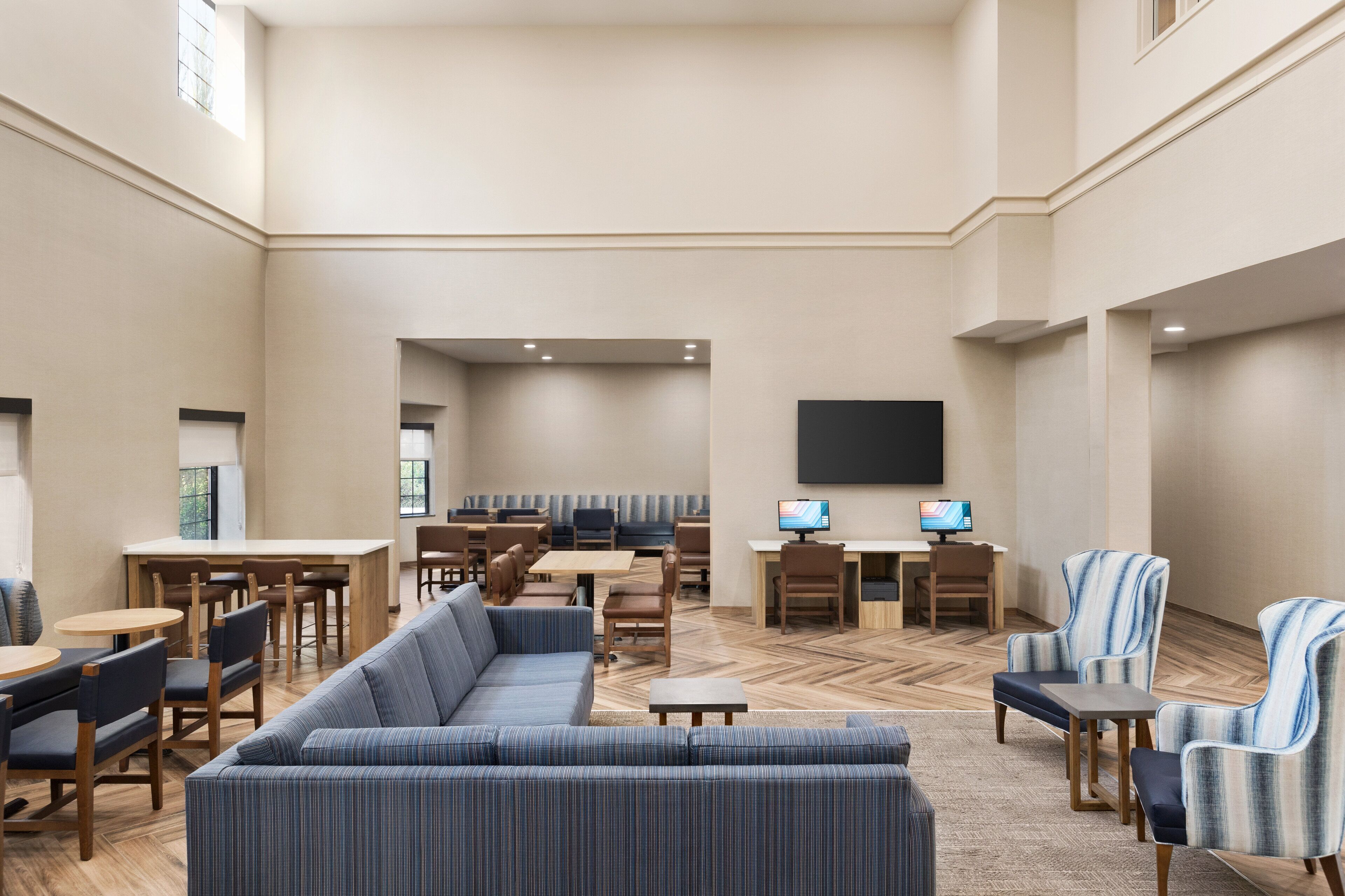 A spacious lobby with a large sofa, dining tables, and chairs, a flat-screen TV, and a herringbone-patterned floor.