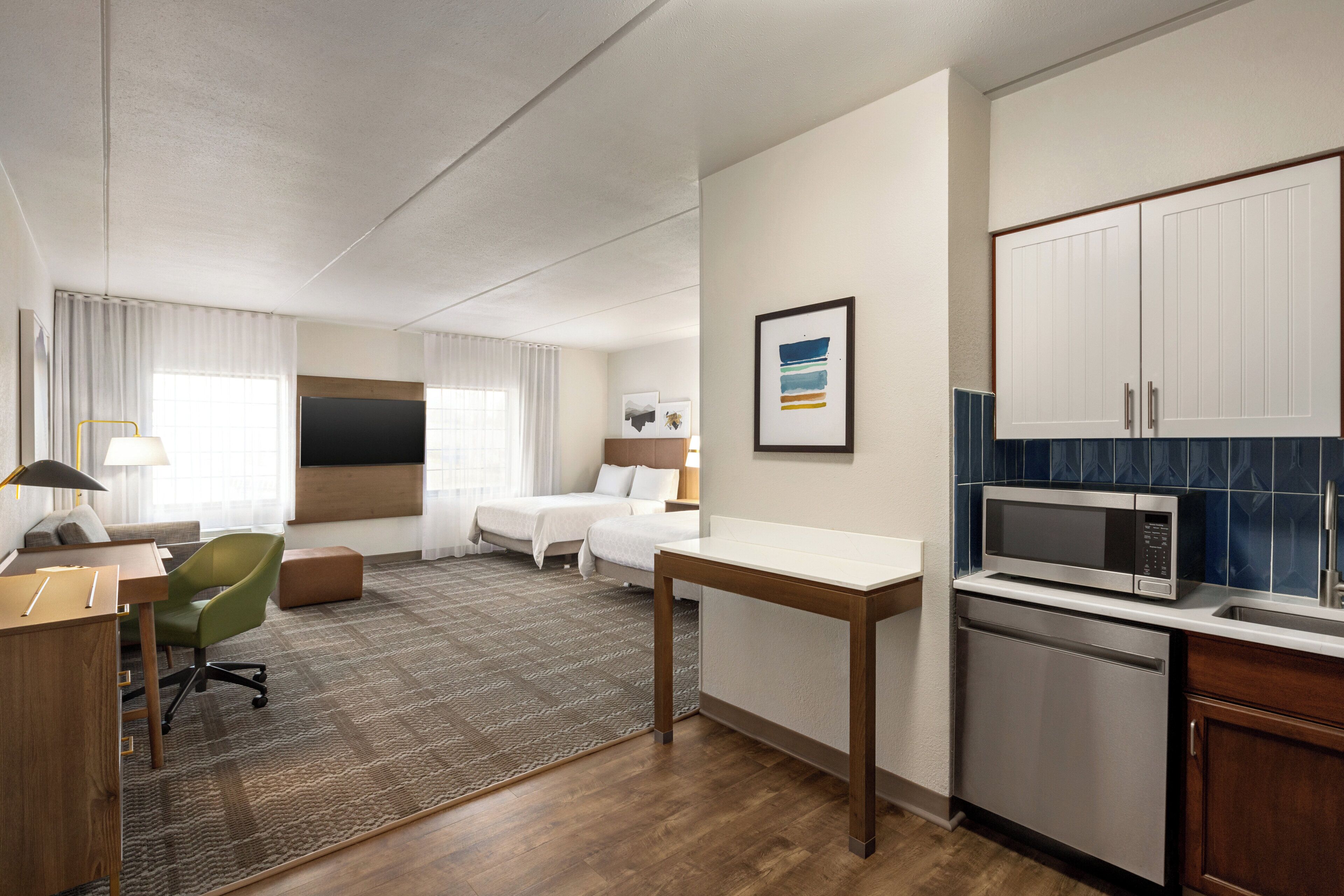 A hotel room with a bed, a desk, a chair, a small kitchenette, and a bathroom.