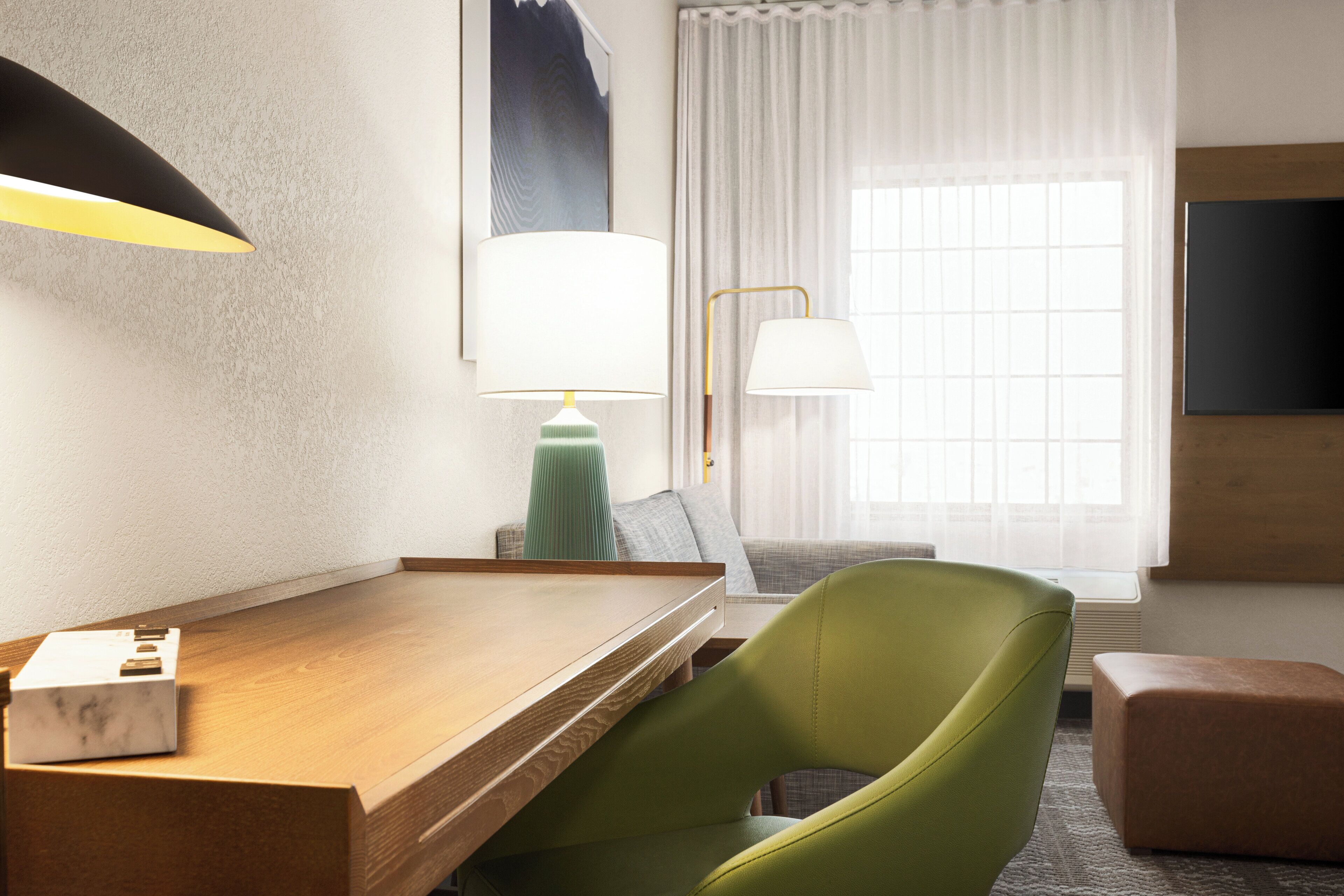 A hotel room with a wooden desk, a green chair, a lamp, a television, and a window with curtains.