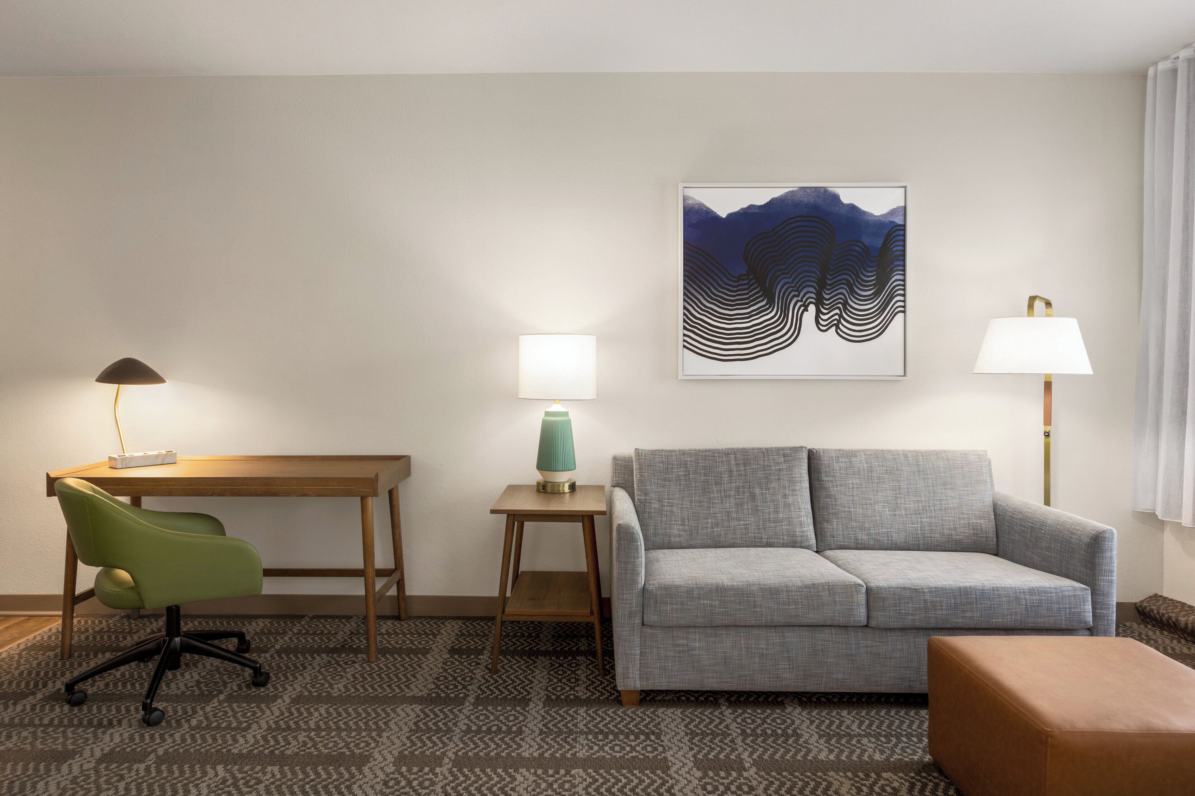 A hotel room with a desk, a green chair, a grey sofa, a patterned carpet, and a wall-mounted artwork.
