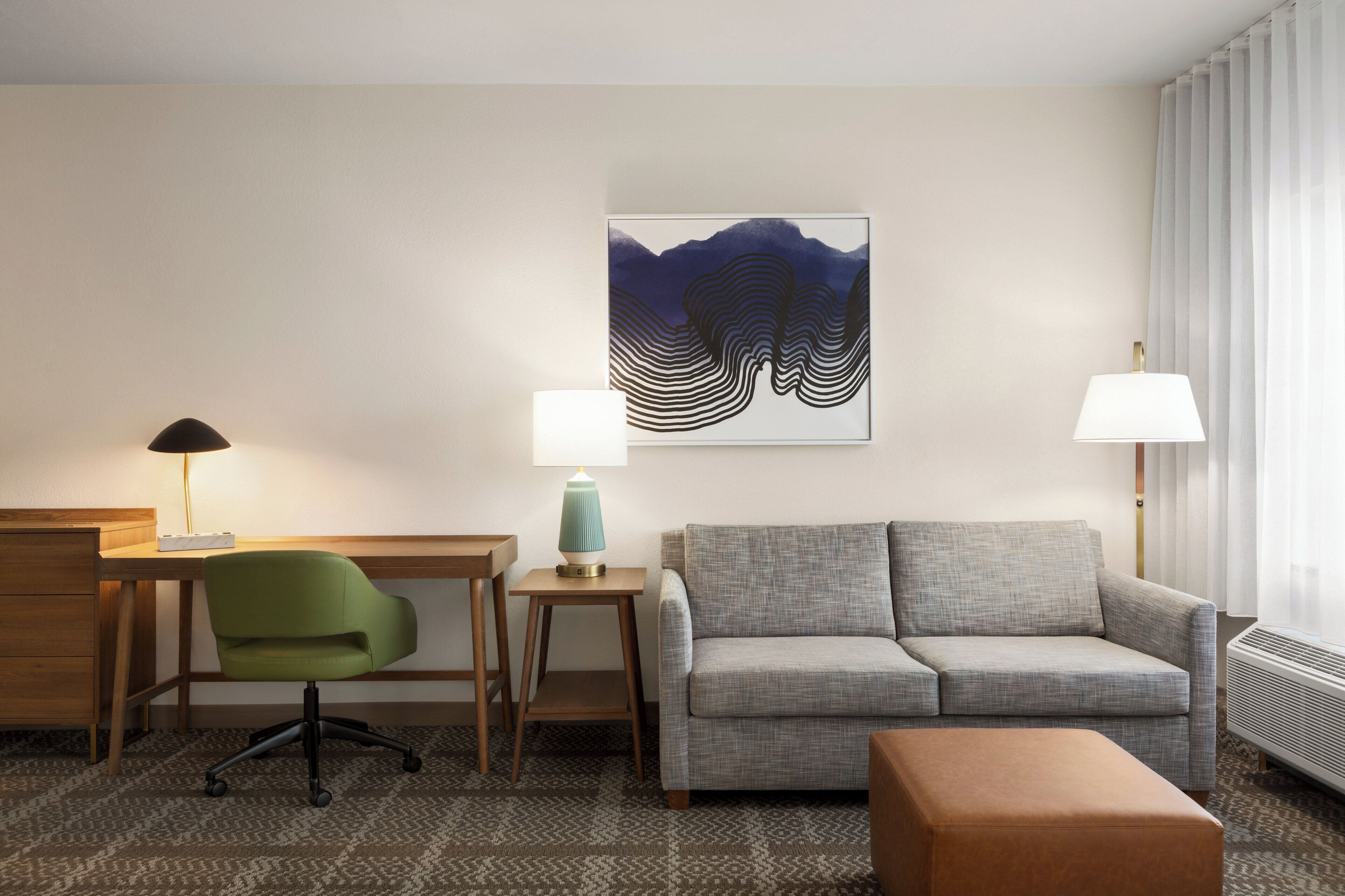 Een hotelkamer met een bureau, een groene bureaustoel, een grijze bank, een klein tafeltje met een lamp en een groot abstract schilderij aan de muur.