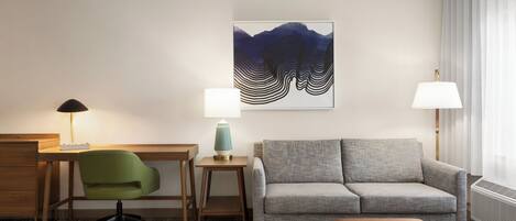 Een hotelkamer met een bureau, een groene bureaustoel, een grijze bank, een klein tafeltje met een lamp en een groot abstract schilderij aan de muur.