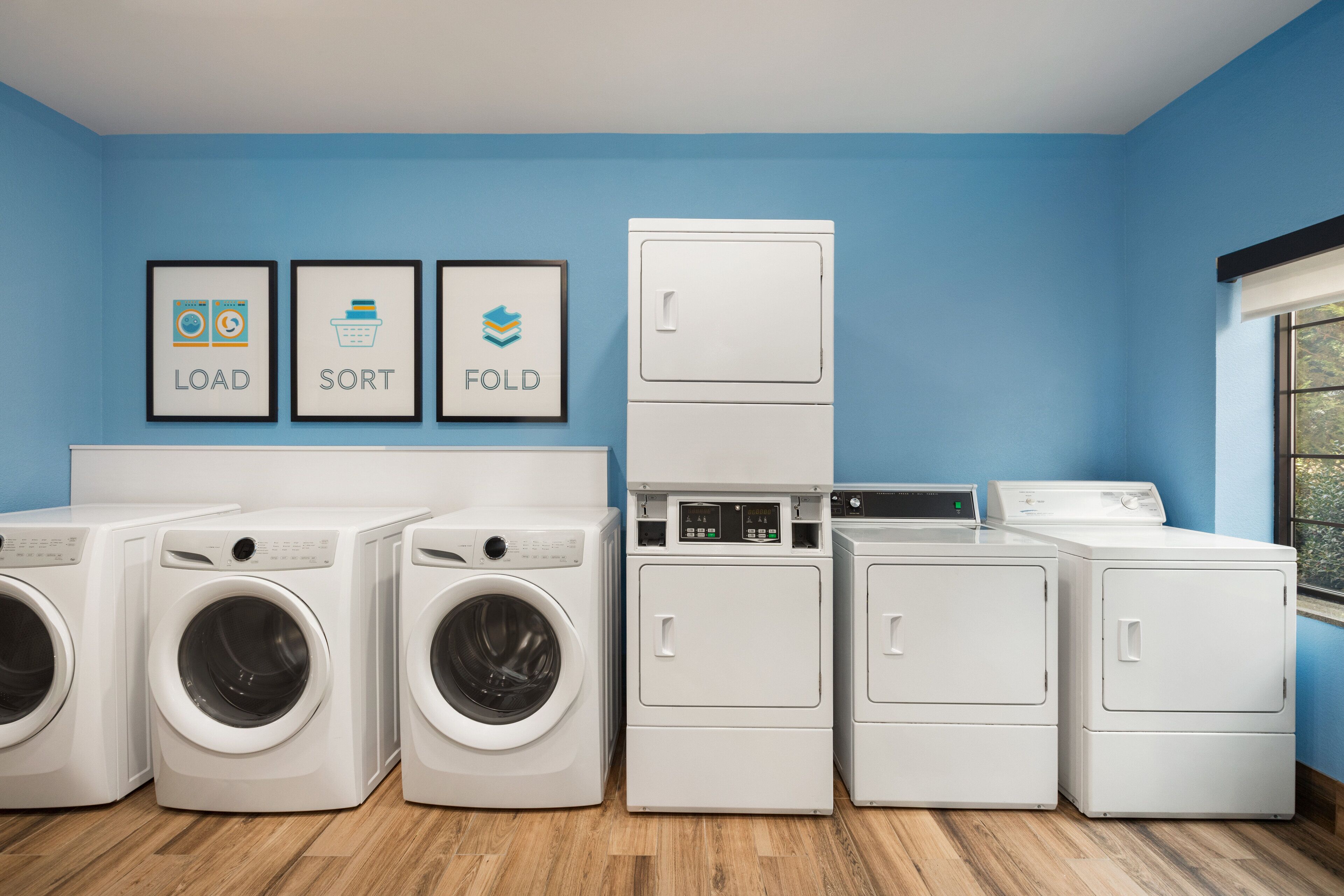 A laundry room with four washing machines, a dryer, and three framed signs labeled "LOAD," "SORT," and "FOLD."
