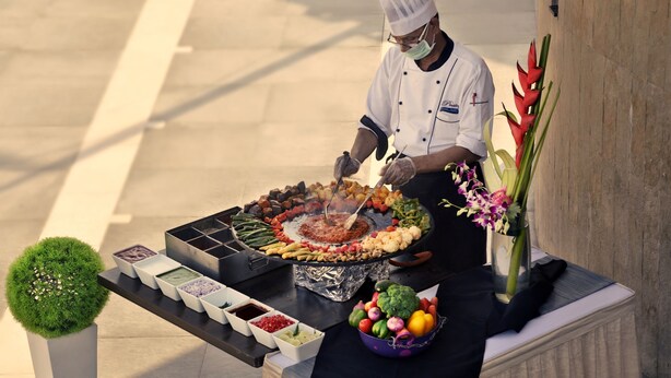 Un chef preparando comida al aire libre con una parrilla, verduras y salsas en una mesa.