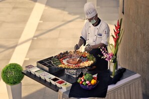Een kok bereidt eten buiten, met een barbecue, groenten en kruiden op een tafel.