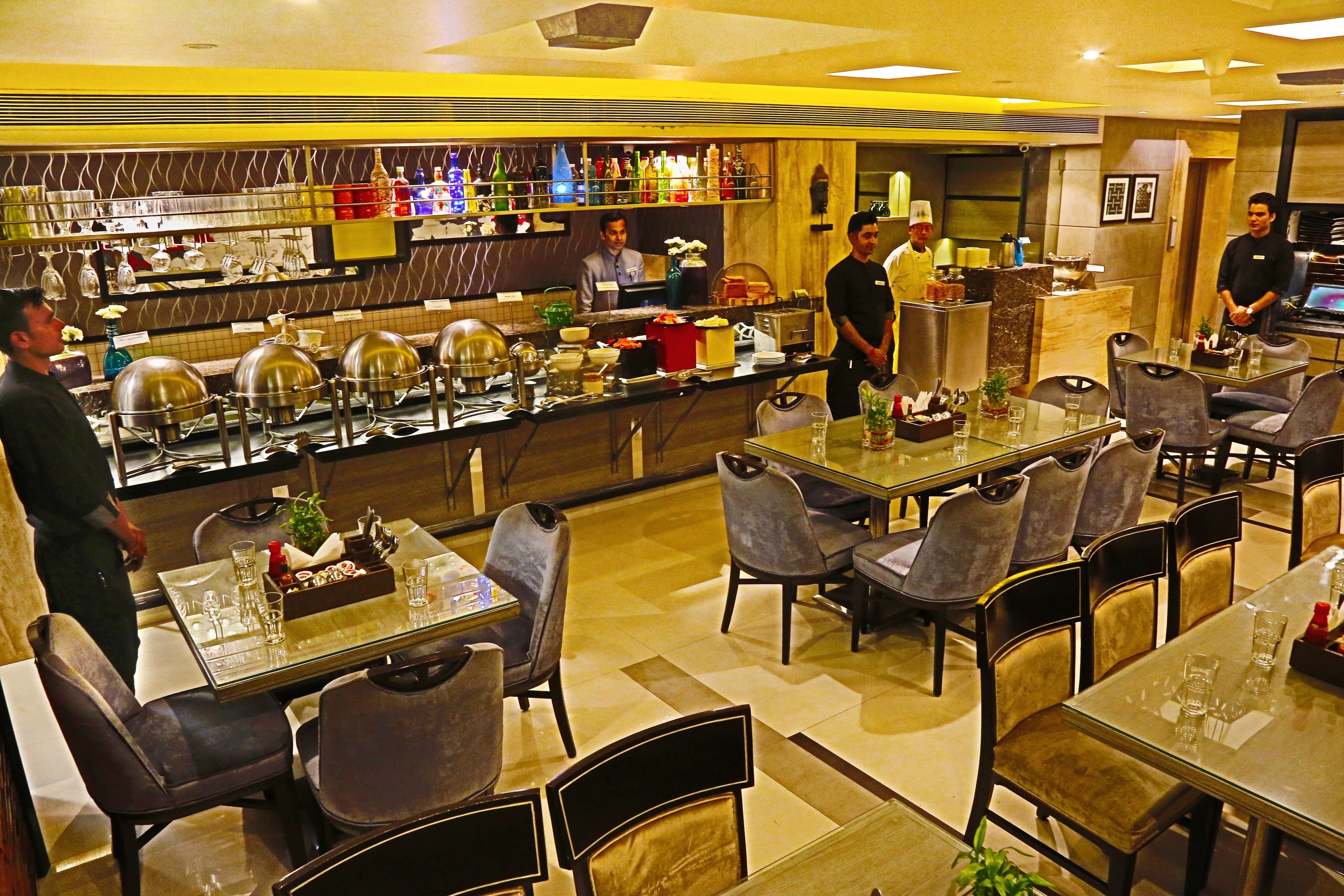 Restaurante com várias mesas e cadeiras, área de buffet com comida e bebidas, e cozinha ao fundo.