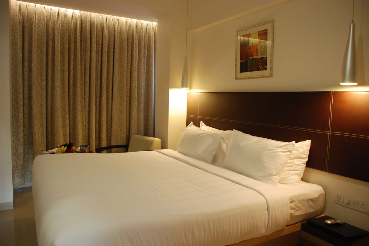 Habitación de hotel con una cama grande, cabecero, mesita de noche, lámpara, una silla y un cuadro en la pared.